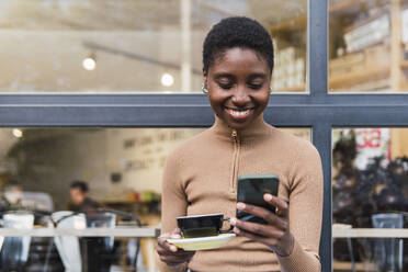 Glückliche junge Frau benutzt ihr Smartphone vor einem Café - PNAF05779
