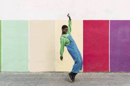 Happy woman standing on tiptoes in front of multi colored wall - PNAF05786