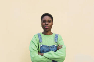 Playful young woman sticking out tongue and standing with arms crossed in front of beige wall - PNAF05792