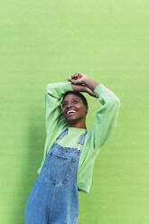 Happy young woman with arms raised standing in front of green wall - PNAF05794
