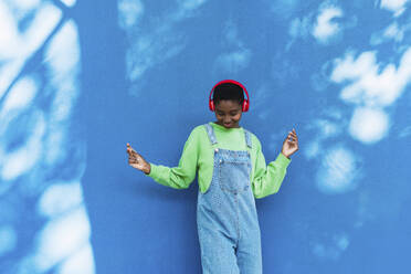 Happy woman listening to music through wireless headphones and dancing in front of blue wall - PNAF05798
