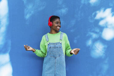 Glückliche junge Frau, die über drahtlose Kopfhörer vor einer blauen Wand Musik hört - PNAF05800