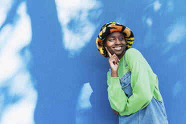 Glückliche junge Frau mit buntem Hut, die ihr Gesicht vor einer blauen Wand berührt - PNAF05807