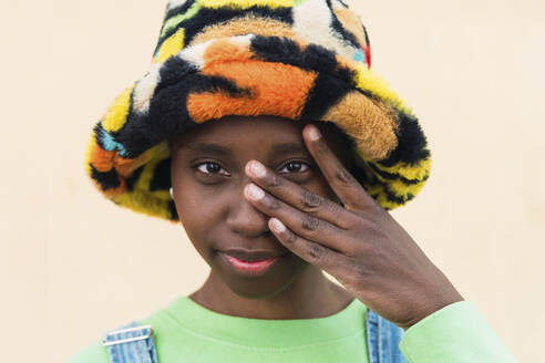Junge Frau mit mehrfarbigem Hut und berührendem Gesicht vor beigem Hintergrund - PNAF05811