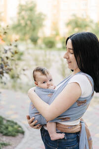 Lächelnde Mutter hält ihre Tochter im Tragetuch im Park - SSYF00169
