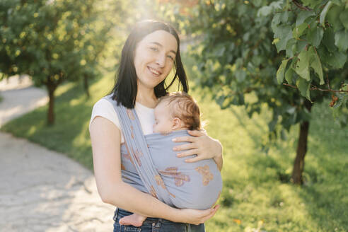 Lächelnde Mutter, die ihre schlafende Tochter in einer Babytrage im Park trägt - SSYF00174