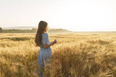 Mädchen mit langen Haaren steht im Weizenfeld bei Sonnenaufgang - AAZF00782
