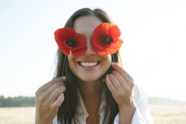Lächelnde Frau hält sich rote Mohnblumen vor die Augen - AAZF00844