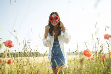 Smiling woman holding red poppies in front of eyes at field - AAZF00845