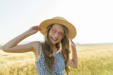 Lächelndes Mädchen mit sternförmiger Sonnenbrille und Hut auf einem Feld - AAZF00852