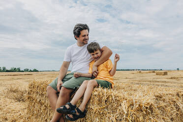 Lächelnder Vater mit Arm um den Sohn, der in einem Weizenfeld sitzt - VSNF01244