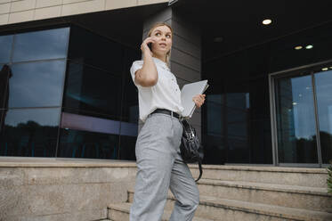 Businesswoman talking on smart phone standing in front of building - ALKF00407