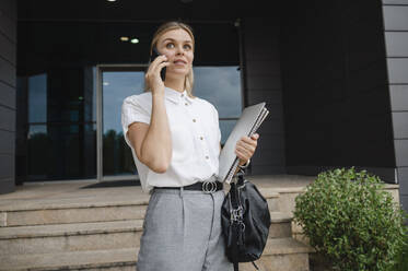 Businesswoman talking on mobile phone standing in front of building - ALKF00408