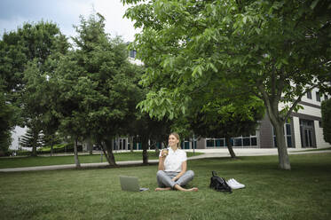 Happy businesswoman sitting on grass with coffee cup and laptop - ALKF00426
