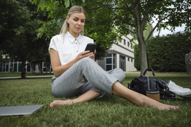 Lächelnde Geschäftsfrau sitzt im Gras und benutzt ihr Smartphone - ALKF00434