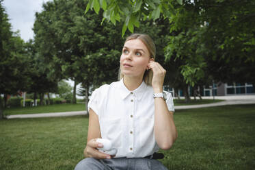 Geschäftsfrau mit drahtlosen In-Ear-Kopfhörern im Büropark sitzend - ALKF00436