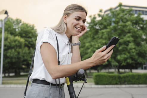 Glückliche Geschäftsfrau, die ihr Smartphone benutzt und sich auf einen Elektroroller stützt - ALKF00444