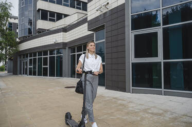 Smiling businesswoman walking with electric push scooter - ALKF00448