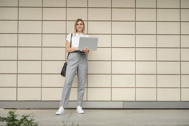 Lächelnde Geschäftsfrau mit Laptop vor einer Wand stehend - ALKF00470