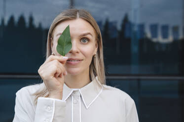 Smiling blond businesswoman holding leaf over eye - ALKF00482