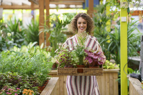 Lächelnde Frau mit einem Tablett voller Topfpflanzen in einer Gärtnerei - JSMF02812