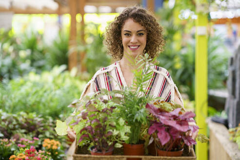 Frau mit lockigem Haar hält Topfpflanzen in einem Tablett in einer Gärtnerei - JSMF02813