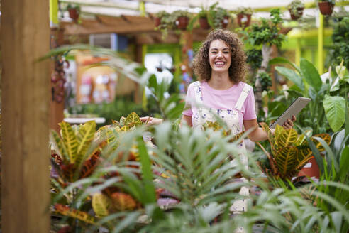 Happy woman holding tablet PC amidst plants in nursery - JSMF02832