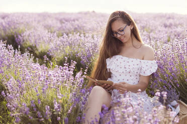 Smiling woman with long hair reading book in lavender field - ONAF00598