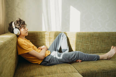 Nachdenklicher Junge mit Kopfhörern, der sich auf dem Sofa entspannt - ANAF01815