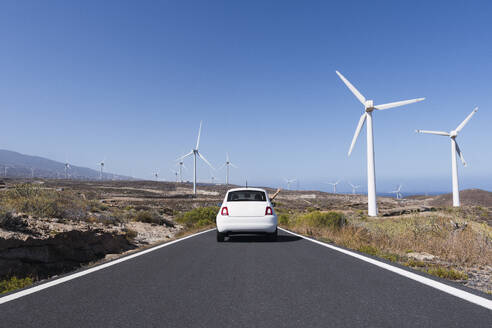 Auto fährt auf der Straße inmitten von Windkraftanlagen unter blauem Himmel - PNAF05854