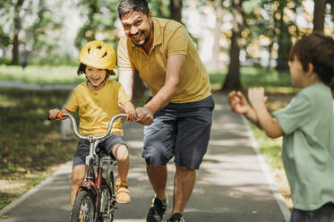 Lächelnder Vater hilft seinem Sohn beim Fahrradfahren im Park - ANAF01827