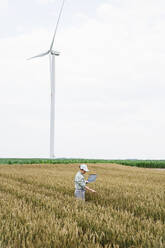 Agronomist examining crops in front of wind turbine at field - EKGF00349