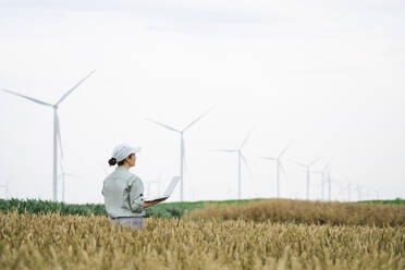Agronomist examining field and standing with laptop in front of wind turbines - EKGF00354
