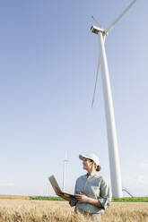 Happy farmer with laptop standing in front of wind turbines on sunny day - EKGF00385