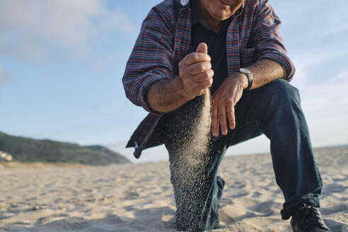 Senior man playing with sand at beach on sunny day - ASGF04083