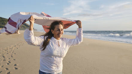 Glückliche ältere Frau mit Kopftuch am Strand an einem sonnigen Tag - ASGF04102