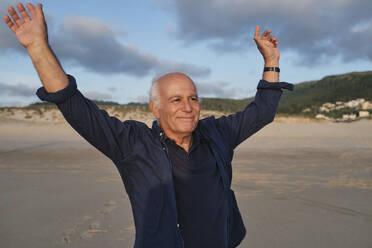 Smiling senior man with arms raised standing at beach - ASGF04116