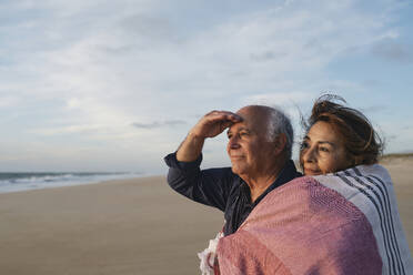 Lächelnder älterer Mann und Frau in Schal eingewickelt am Strand - ASGF04125