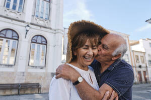 Fröhlicher älterer Mann, der eine Frau mit Hut vor einem Gebäude umarmt - ASGF04178