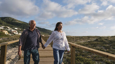 Happy senior couple holding hands walking on boardwalk - ASGF04204