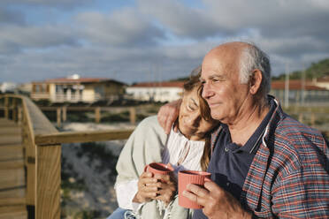 Lächelndes älteres Paar, das Kaffeetassen hält und sich an einem sonnigen Tag umarmt - ASGF04267