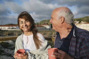 Happy senior woman and man holding coffee cups under cloudy sky - ASGF04268