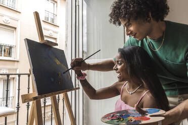 Smiling boyfriend helping girlfriend painting on canvas at home - PBTF00076