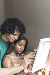 Happy boyfriend helping girlfriend to paint on canvas at home - PBTF00080