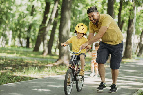 Glücklicher Vater, der seinem Sohn beim Fahrradfahren auf dem Fußweg im Park hilft - ANAF01829