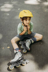 Sad boy wearing helmet sitting on footpath with inline skates - ANAF01832