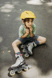 Trauriger Junge mit Helm, der mit Inline-Skates auf dem Gehweg sitzt - ANAF01832