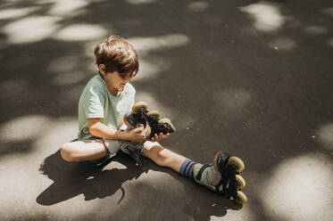 Boy removing inline skates sitting on footpath - ANAF01833