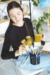 Kontemplativer Künstler, der auf einem Stuhl im Atelier sitzend den Pinsel auswählt - YHF00003