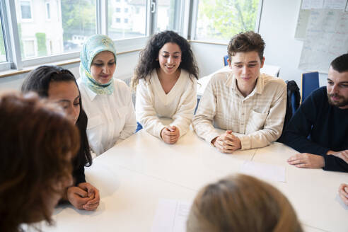 Schüler sitzen mit verschränkten Händen um einen Tisch im Klassenzimmer - NJAF00453